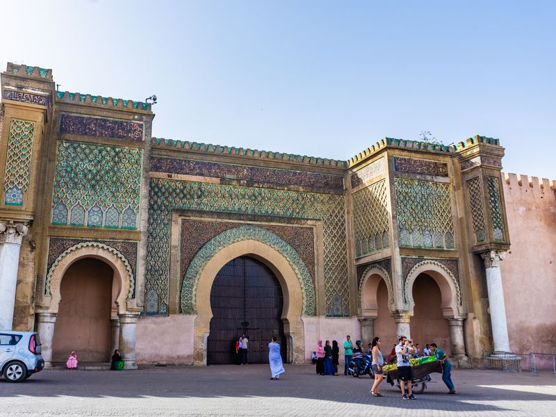 The ornate Bab Bou Jeloud gate in Fes, Morocco, with its intricate blue and green tile work. People are present near the gate, giving context to its size and showcasing the vibrant cultural scene.