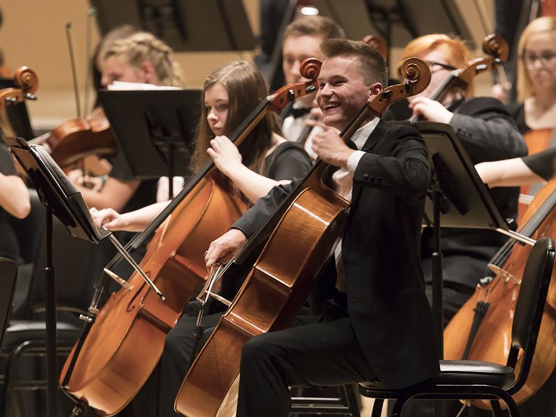 Young adult cellist smiling while performing with the orchestra.