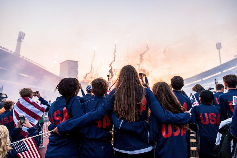 Group of young people in USA jackets, arms around each other, watching fireworks in a stadium.