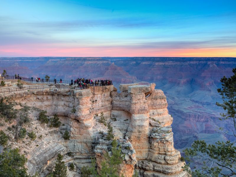 A group of tourists stand on the edge of a cliff overlooking a vast canyon bathed in sunset's orange light, Grand Canyon National Park.