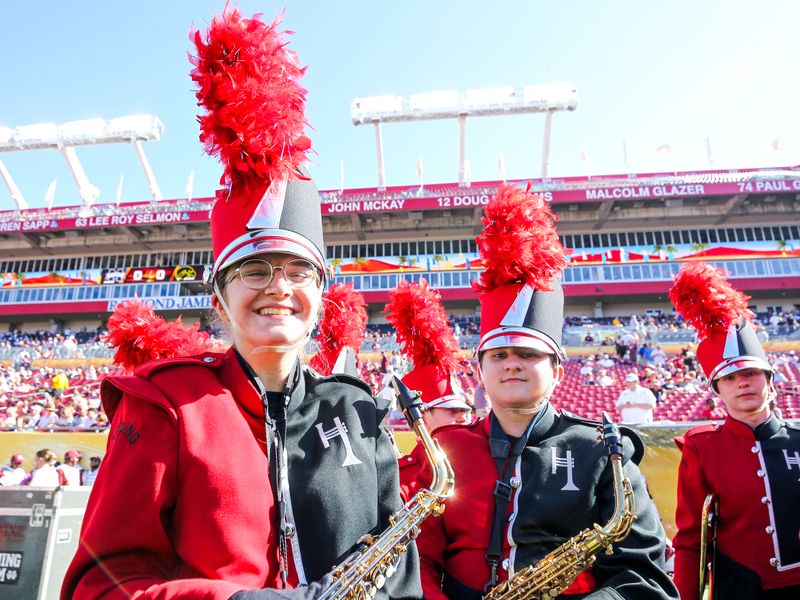 Group of teenage band members smiling in uniform holding saxophones on a football field.