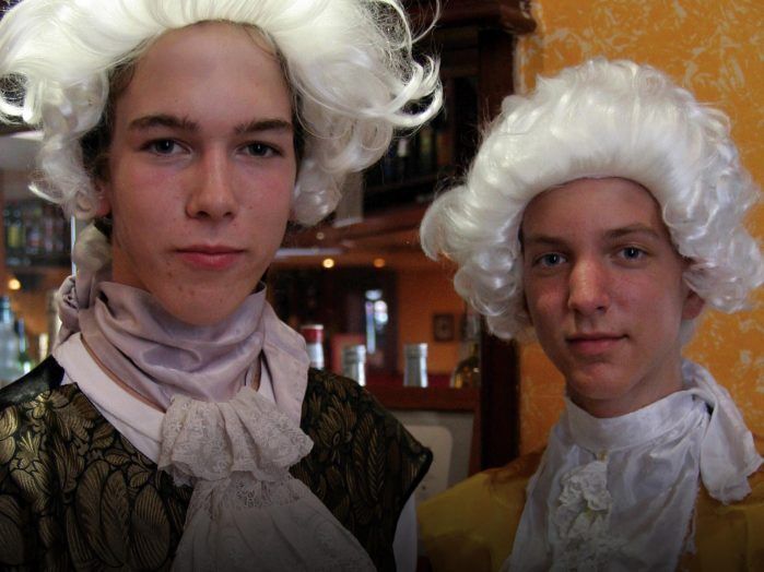 Two teenagers in colonial costumes, complete with white wigs and period attire, pose against the backdrop of a modern-day bar area.