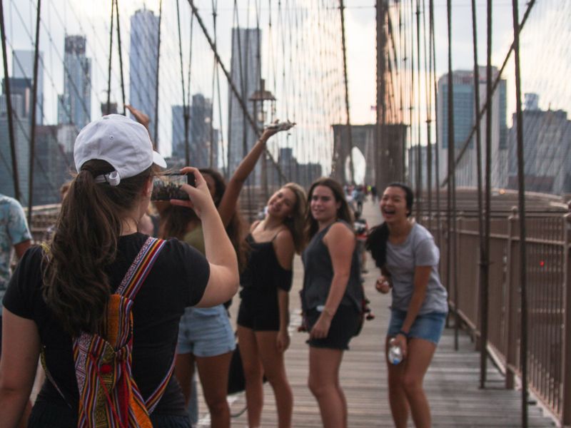 A group of friends taking a selfie on the Brooklyn Bridge with the New York City skyline in the background.