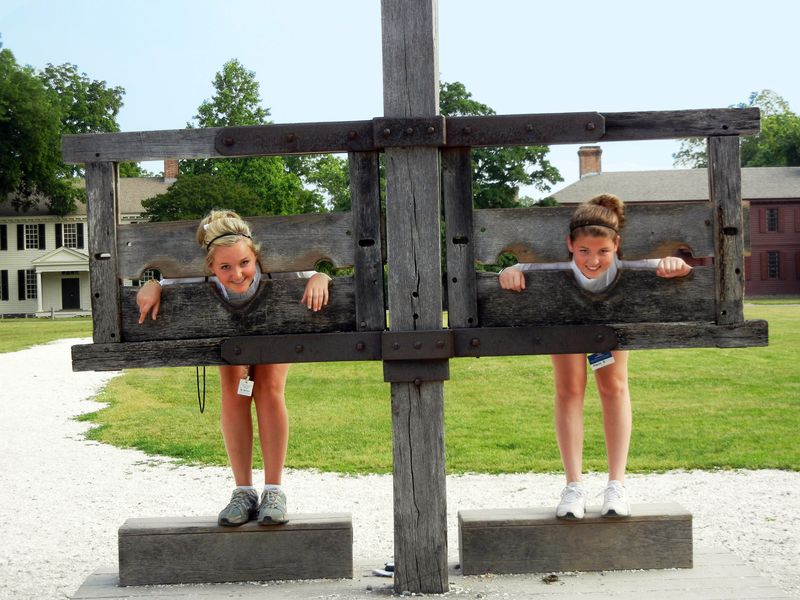 Two teenage girls playfully experience history by placing their heads and hands in a wooden stockade at a colonial village.