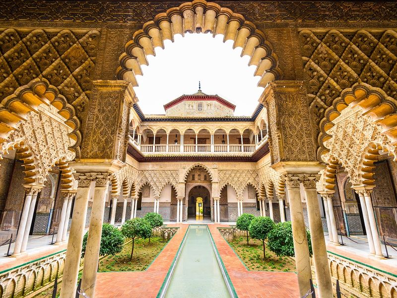 A picturesque view of the courtyard in the Alcázar of Seville, Spain, with its ornate arches, reflecting pool, and vibrant gardens.