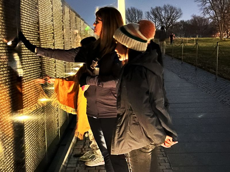 Three people stand at the Vietnam Veterans Memorial in Washington, DC at night, their faces illuminated by the light reflecting off the wall.