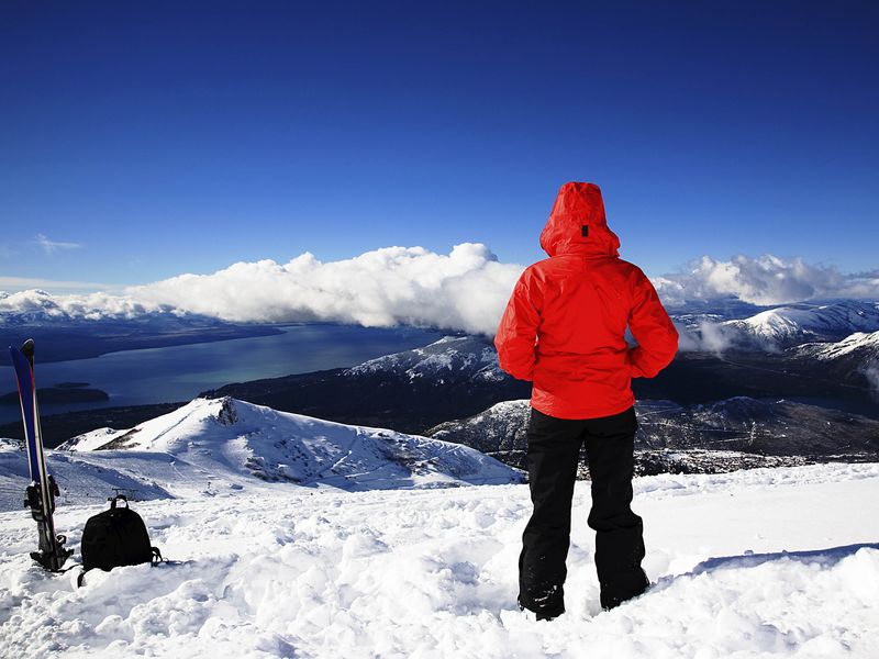 Skier in red jacket enjoying a mountaintop view of a lake and snow-capped peaks.