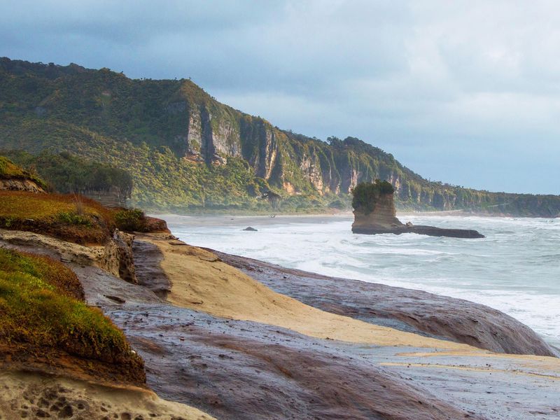 A captivating view of a rugged coastline, with a rocky beach, imposing cliffs, and a unique rock structure in the ocean, under a dynamic sky.