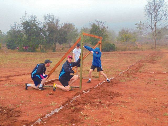 A group of adults building a wooden structure on a red dirt field.