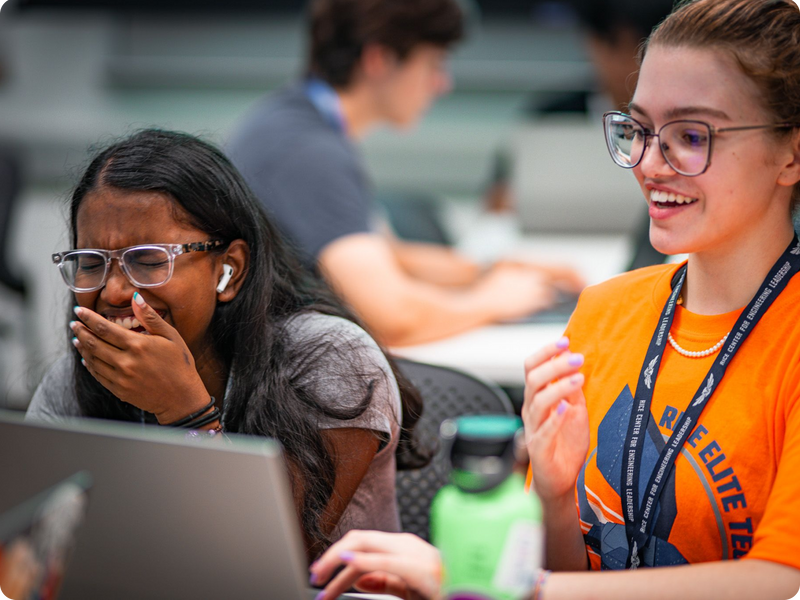 Two teenage girls engaged with a laptop, one laughing and the other smiling.