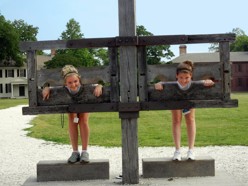 Two teenage girls playfully pose in a pillory at a historical site.