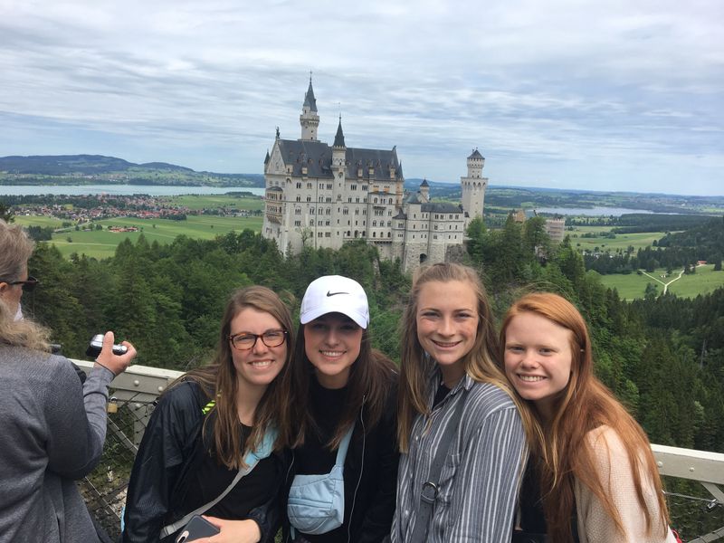 Four young adults take a selfie with the majestic Neuschwanstein Castle in the backdrop.