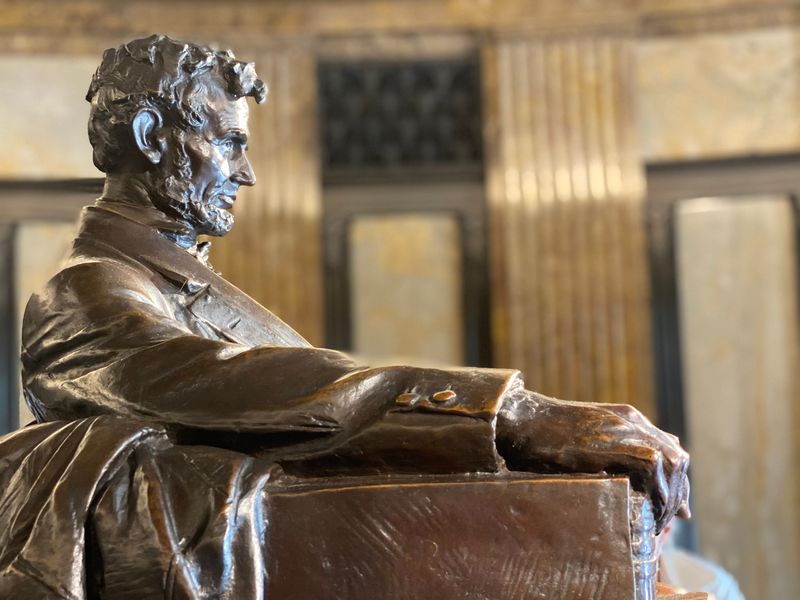 A bronze bust of Abraham Lincoln in profile view, displayed against a softly lit backdrop with columns.