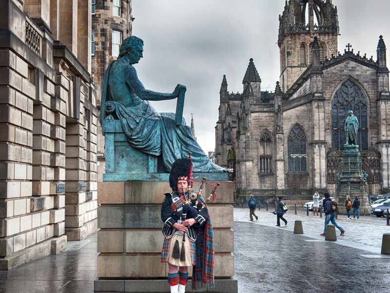 A bagpiper in traditional Scottish attire plays the bagpipes in front of a statue in Parliament Square, Edinburgh, with St. Giles' Cathedral in the background.