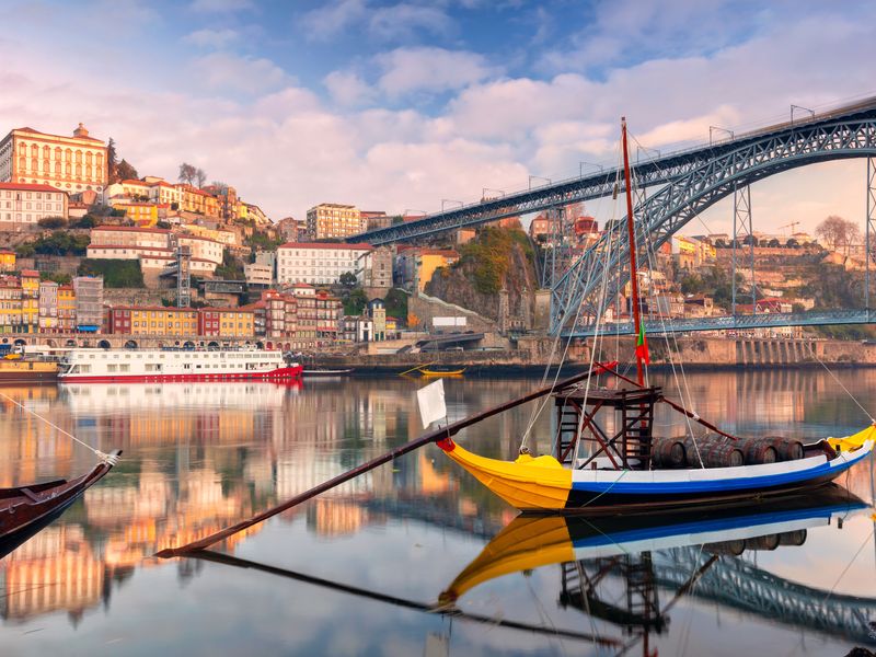 Sunrise over the Douro River in Porto, Portugal, with the Dom Luís I Bridge and a traditional Rabelo boat in the foreground.