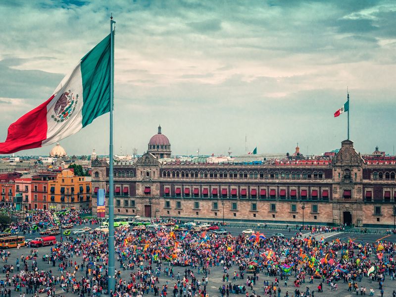 A large crowd gathered in Zócalo, Mexico City, with the Mexican flag prominently displayed and the National Palace in the background.