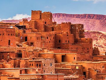 An ancient ksar in Ait Benhaddou, Morocco, made of orange clay bricks, stands tall against a desert mountain backdrop.  