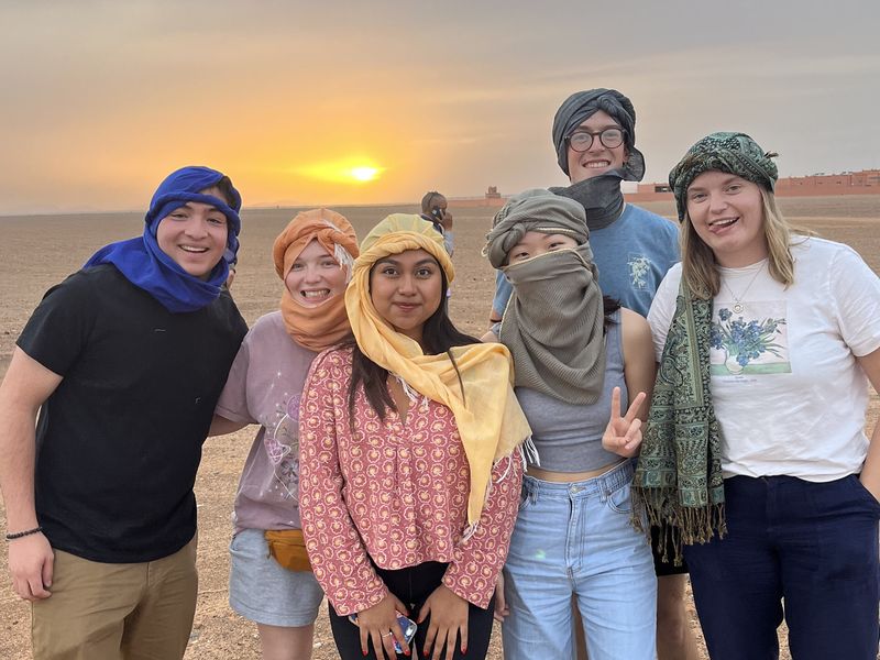 Five young adults wearing traditional desert headwear pose together for a photo in the Sahara Desert at sunset.