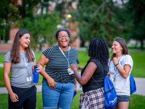 Four young adult women share a laugh together on a green college campus.  They are wearing casual clothes and lanyards, suggesting they may be attending an event.