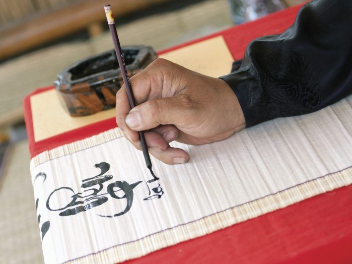 A close-up of a hand practicing calligraphy on a white scroll, using a traditional brush and black ink.