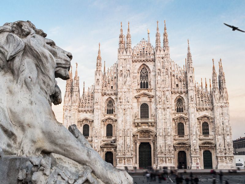 The Duomo di Milano in Milan, Italy. The foreground showcases a majestic marble lion statue, adding depth to the image. A flock of birds soars gracefully across the backdrop of the towering cathedral, bathed in the warm hues of the setting sun.