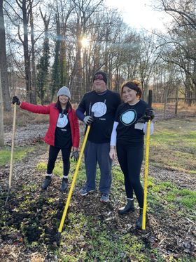 Three young adults volunteering in a community garden, using pitchforks and wearing matching t-shirts.