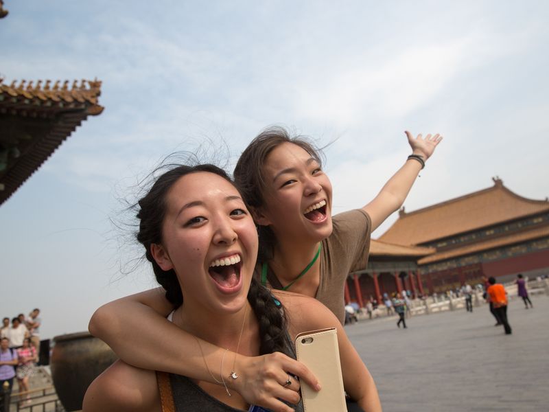 Two young women smile and laugh together in front of a large temple in the Forbidden City, Beijing, China.