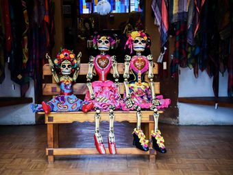 Three intricately dressed Catrina dolls sit on a bench, their colorful attire contrasting with their skeletal frames.