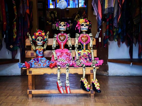 Three intricately dressed Catrina dolls sit on a bench, their colorful attire contrasting with their skeletal frames.