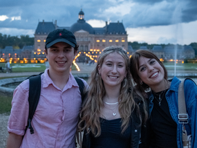 Three young adults smiling for a photo, with a large chateau lit up in the background.