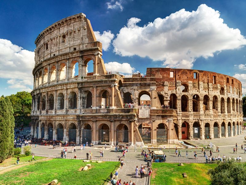 The Colosseum in Rome, Italy on a sunny day with blue sky and white clouds and tourists walking around the perimeter.