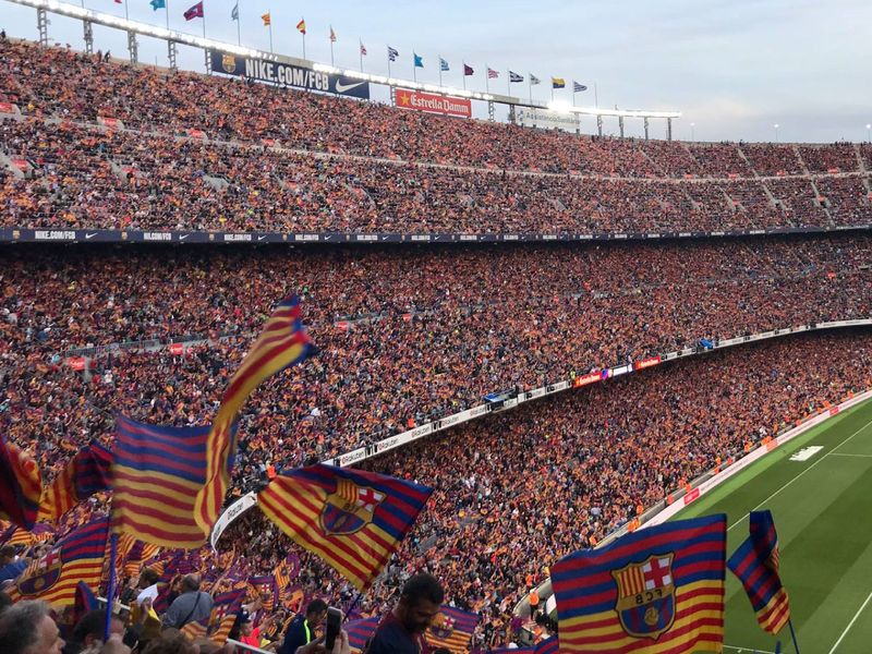 A large crowd of people are watching a soccer game or football match at Camp Nou stadium in Barcelona, Spain. The crowd is mostly wearing orange and waving team flags.