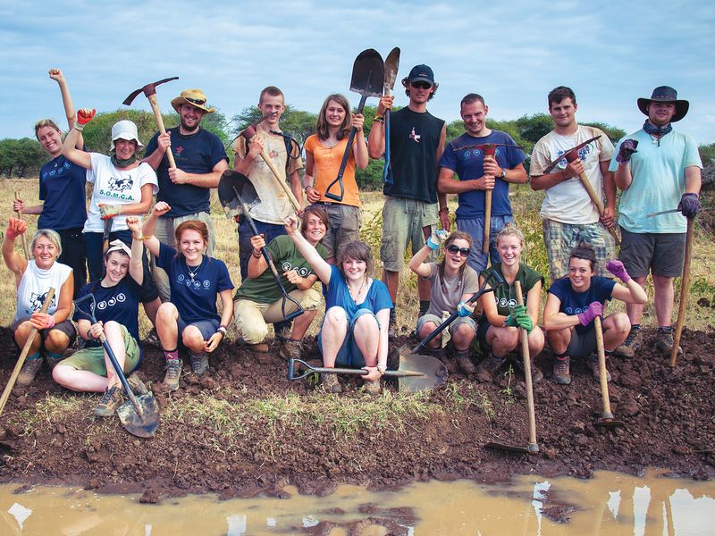 A group of young adult volunteers posing with tools after completing a community service project in a rural, outdoor setting.