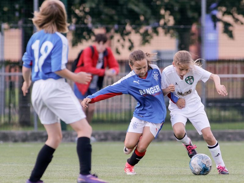 Two young girls compete for the ball during a children's soccer game. The intensity is palpable as one girl in a blue Rangers jersey challenges a girl in a white Hibernian jersey.