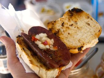 A hand holding a grilled sausage sandwich with onions and peppers.