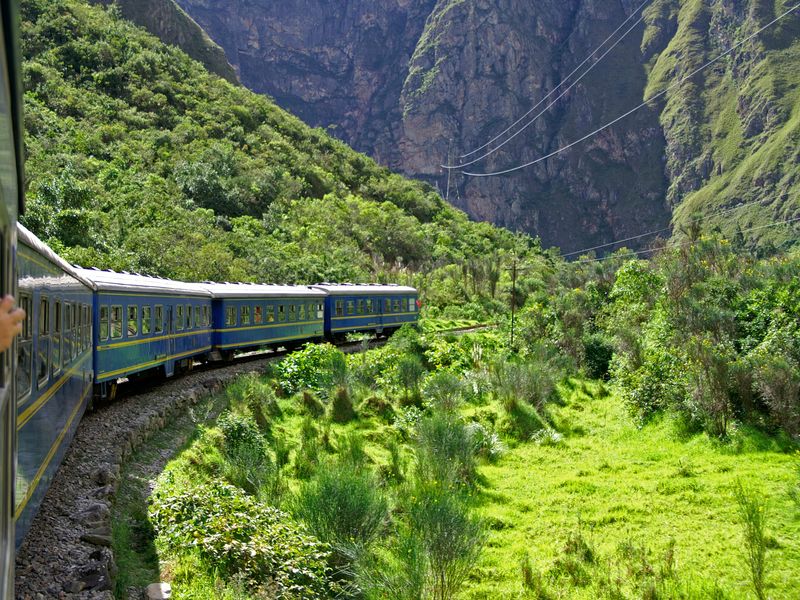 View from a train window of a blue train traveling through a green valley between mountains in the Sacred Valley, Peru.