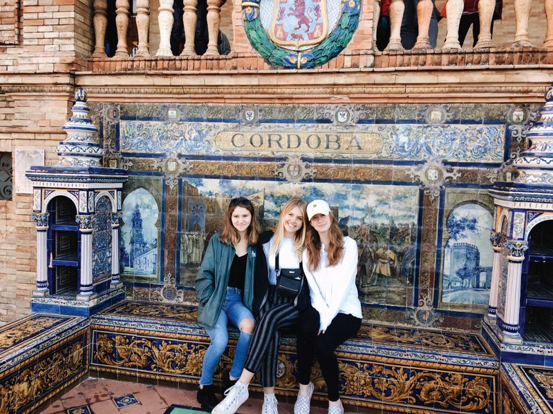 Three young women sit on a bench in front of a wall decorated with Spanish tiles in Seville, Spain. The tiles depict scenes of Cordoba.