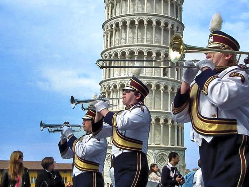 A marching band in white and blue uniforms plays in front of the Leaning Tower of Pisa.