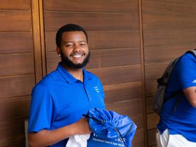 A young man smiles at the camera, holding a drawstring bag and towel. He wears a blue polo shirt and has a beard. The background is a brown wall with horizontal slats.