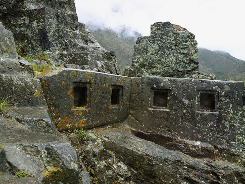 Ancient stone ruins with square openings, set in a rocky landscape under an overcast sky.