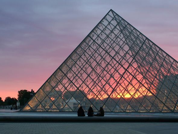 The Louvre Pyramid illuminated by the warm glow of the setting sun, with a few people sitting in front.