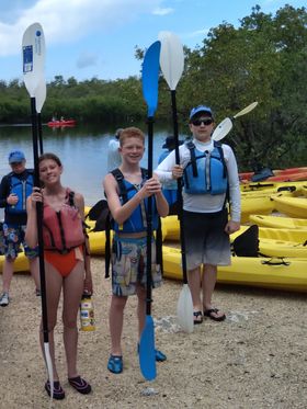 Three young kayakers stand on the beach with their paddles and life jackets, ready for a day on the water.