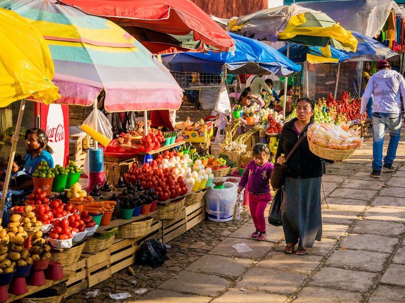 A bustling outdoor market with colorful umbrellas, vendors selling fresh produce, and people shopping.