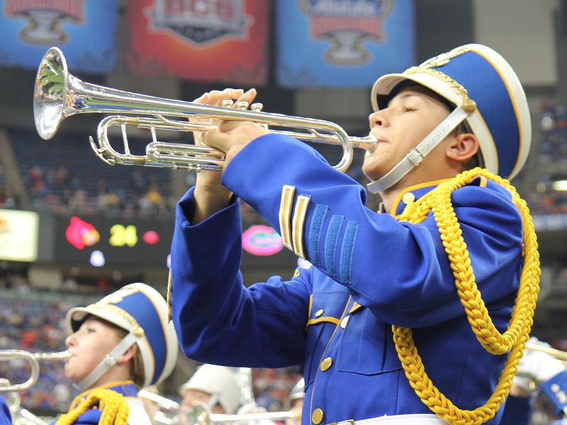 A teenage trumpet player in a blue and yellow uniform performs with a marching band in a stadium at Sugar Bowl in New Orleans.