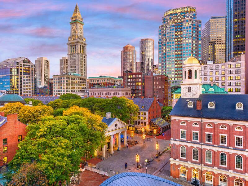 Elevated cityscape view of Boston, MA, featuring Faneuil Hall Marketplace, skyscrapers, trees in autumn colors, lit up during sunset.