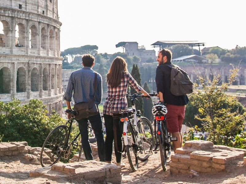 Three young adults take a break from their bike ride touring Rome, Italy to look at the Colosseum.