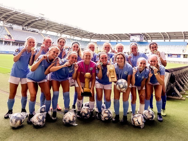 A women's soccer team celebrates winning a championship, holding their trophy and gold medals on the field.