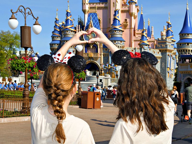 Two teenagers wearing mouse ears stand with their back to the camera and make a heart shape with their hands in front of Cinderella's Castle in Disney World.