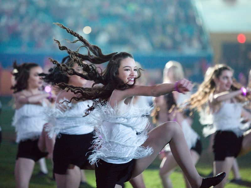 A teenage girl in a white feathered top and black shorts dances energetically on a field as part of a larger group performance. Her long brown hair flows dramatically around her as she kicks high, capturing the dynamism and excitement of the event.