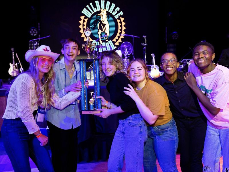 A group of teenagers celebrating their win at a music competition with a trophy. The Wildhorse Saloon sign and stage are visible in the background.